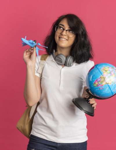 VSMR Visas young pleased pretty caucasian schoolgirl with headphones on neck wearing glasses and back bag holds plane and globe looking up isolated on pink background with copy space
